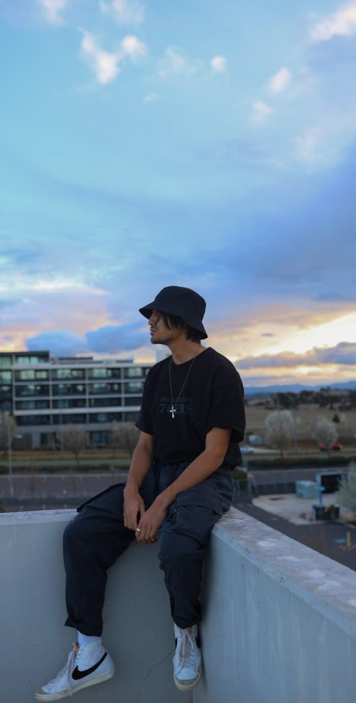 A young man in a bucket hat sits on a rooftop ledge, gazing at the sunset skyline.